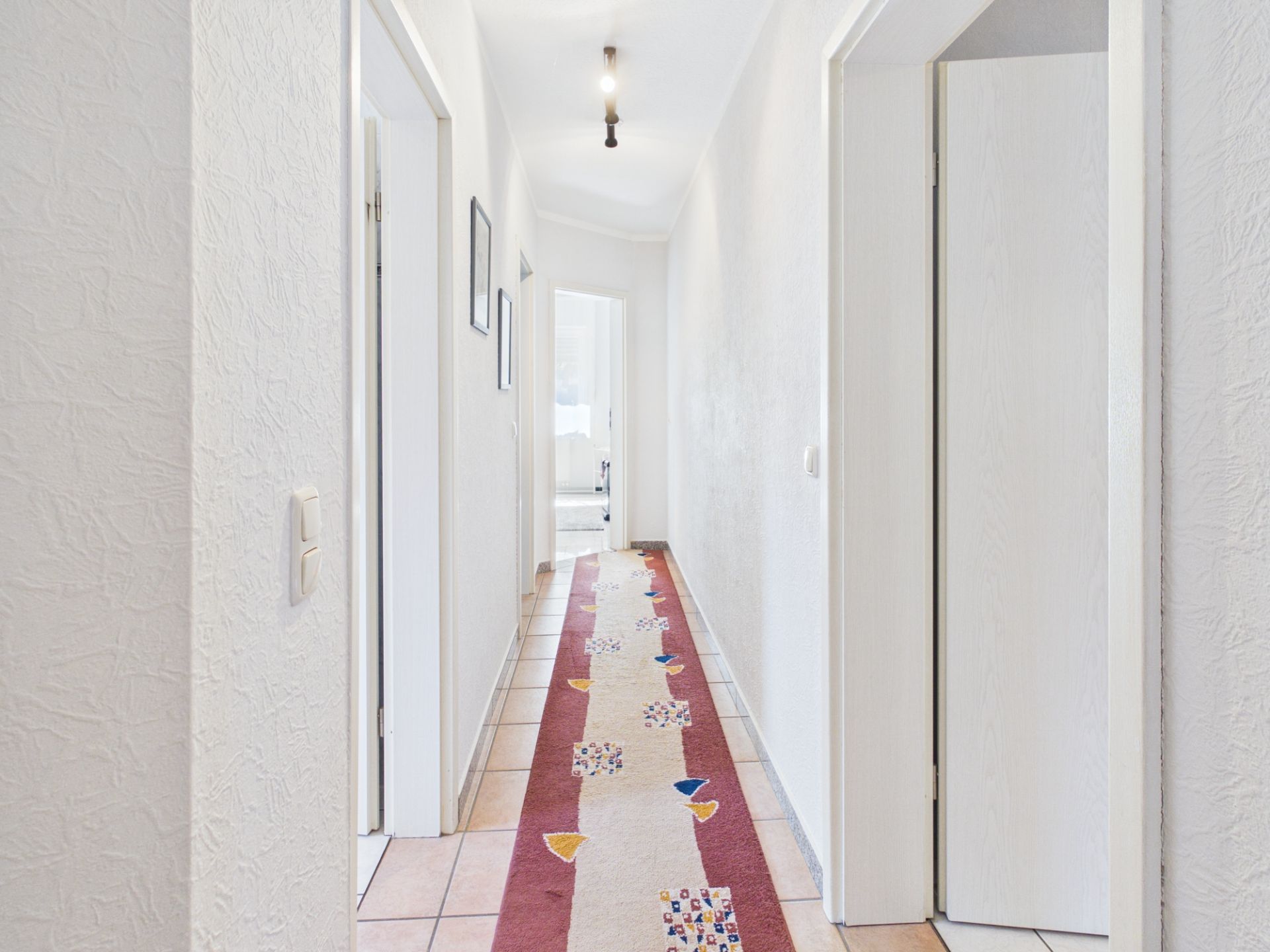 Heller Flur mit buntem Läufer und mehreren offenen Zimmertüren in einer Wohnung.