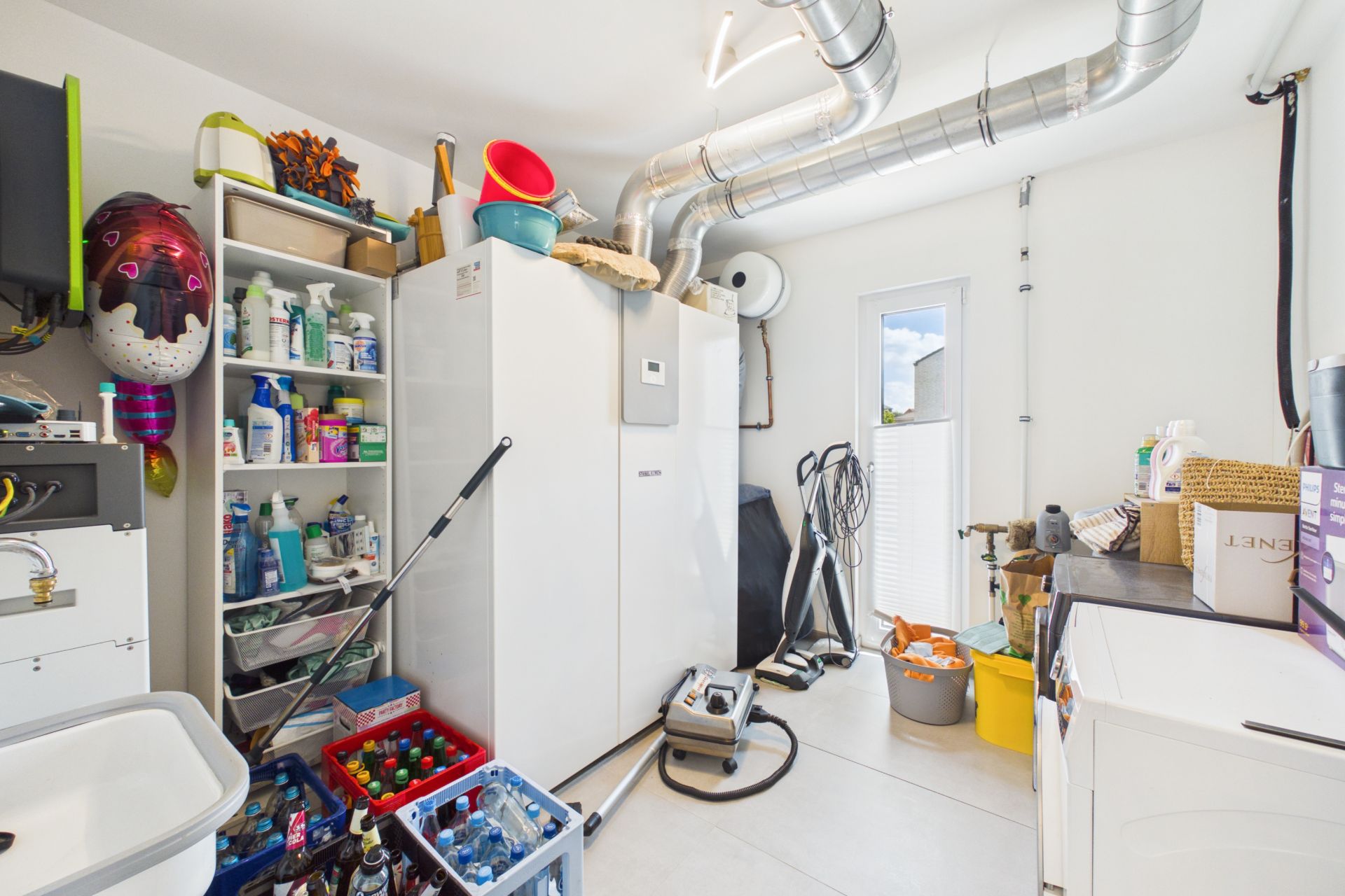 Hauswirtschaftsraum mit Regalen, Reinigungsmitteln, Waschmaschine und großen Lüftungsrohren.