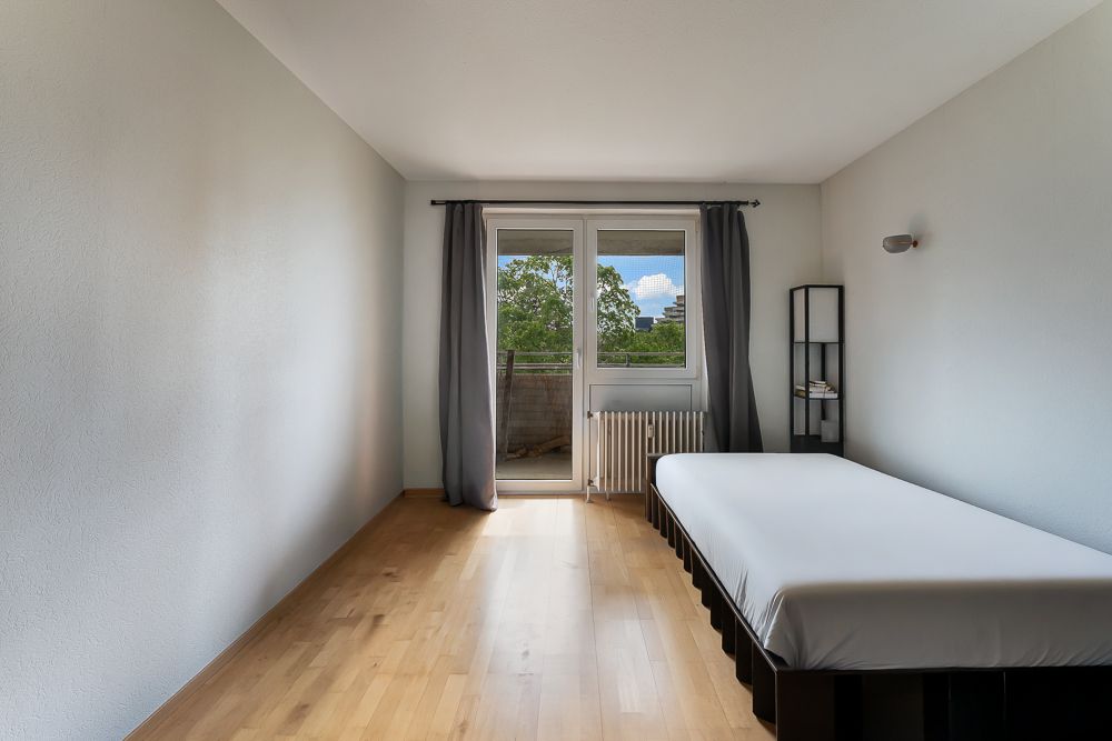 Helles Schlafzimmer mit Einzelbett, Holzfußboden und Fenster mit Blick auf einen Balkon.