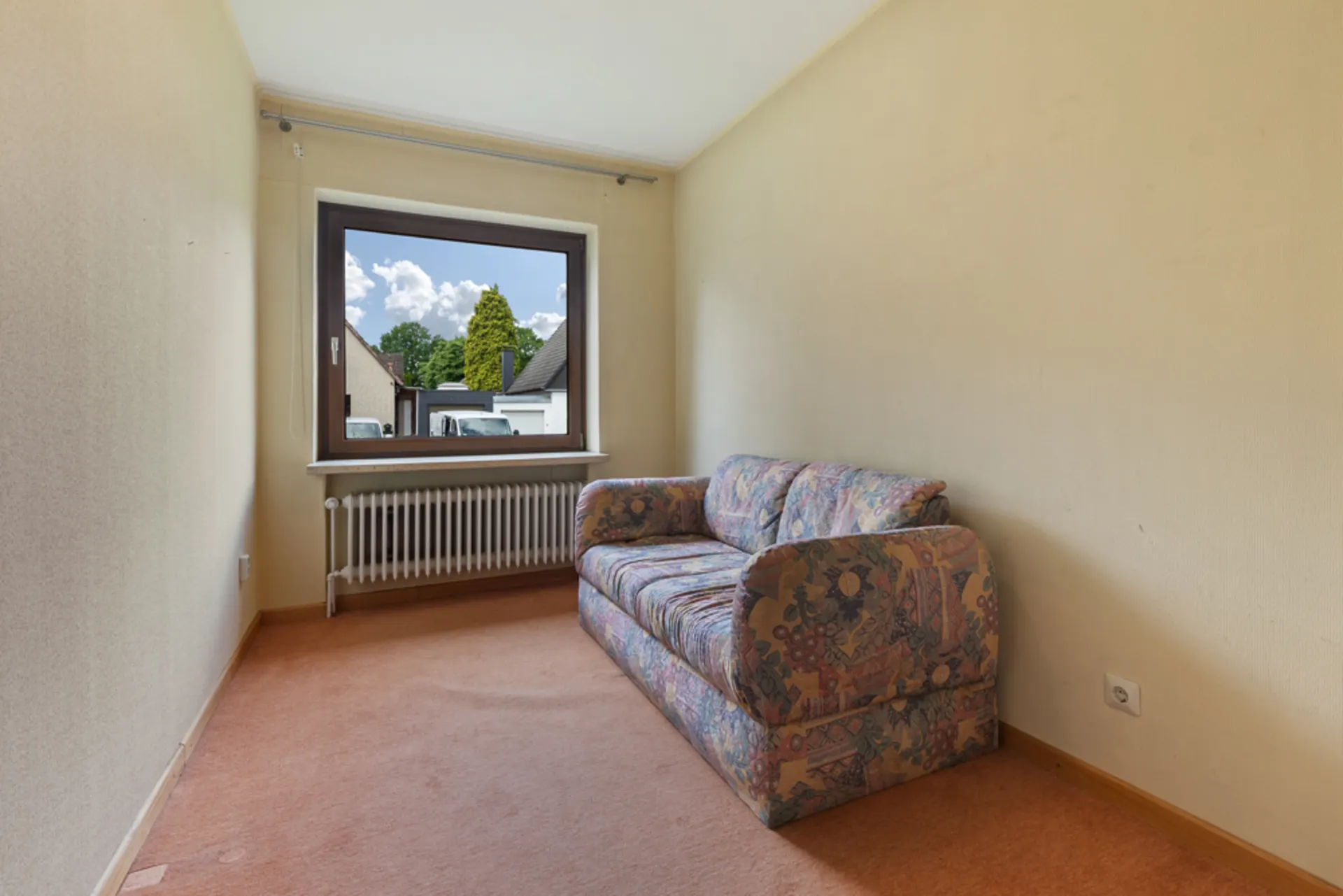 Kleines Zimmer mit gemusterter Couch, Fenster und Heizkörper, beige Wände und Teppichboden.