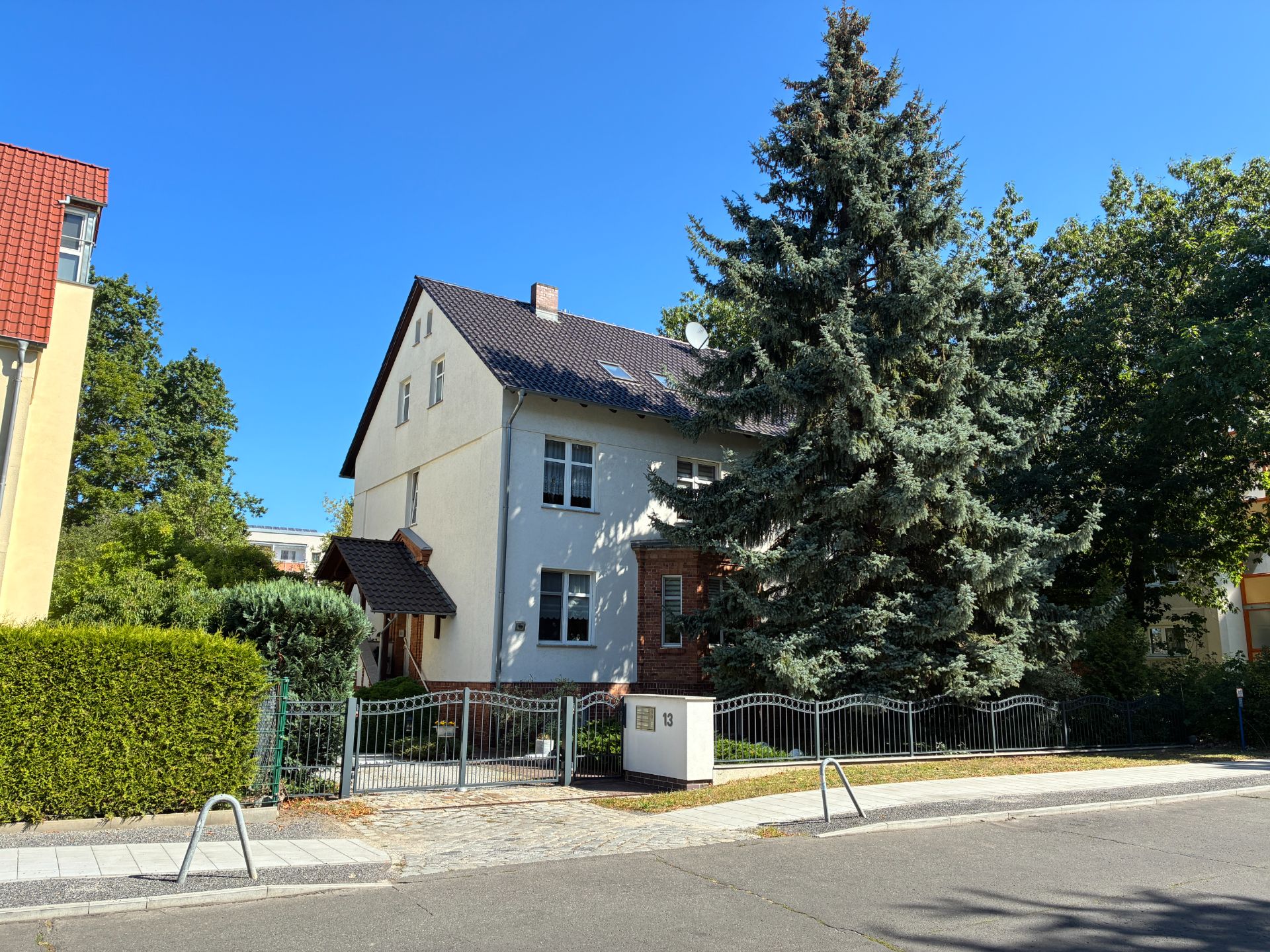 Zweistöckiges Haus mit dunklem Dach und Vorgarten, großer Tannenbaum vorne, blauer Himmel.