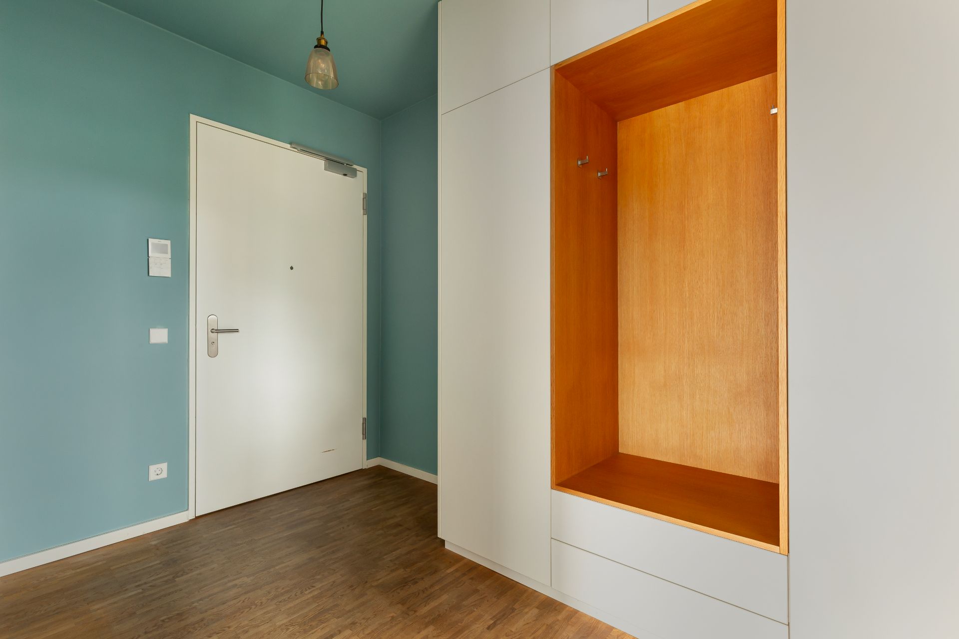 Eingangsbereich mit weißer Tür, blaugrüner Wand und Holzgarderobe.