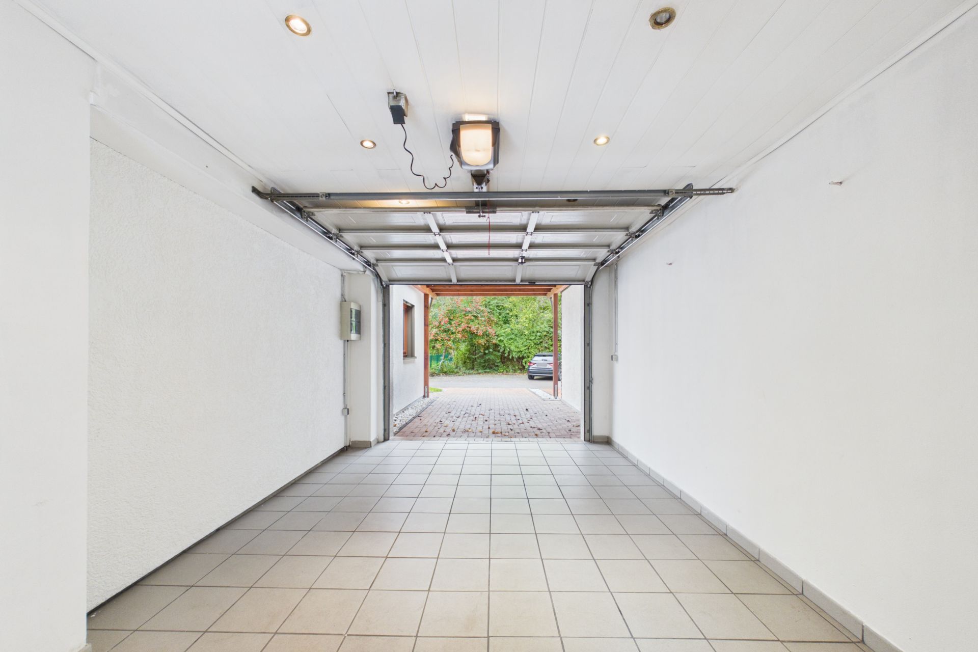 Leere Garage mit weiß gestrichenen Wänden, Fliesenboden und offenem Tor zum Hof.