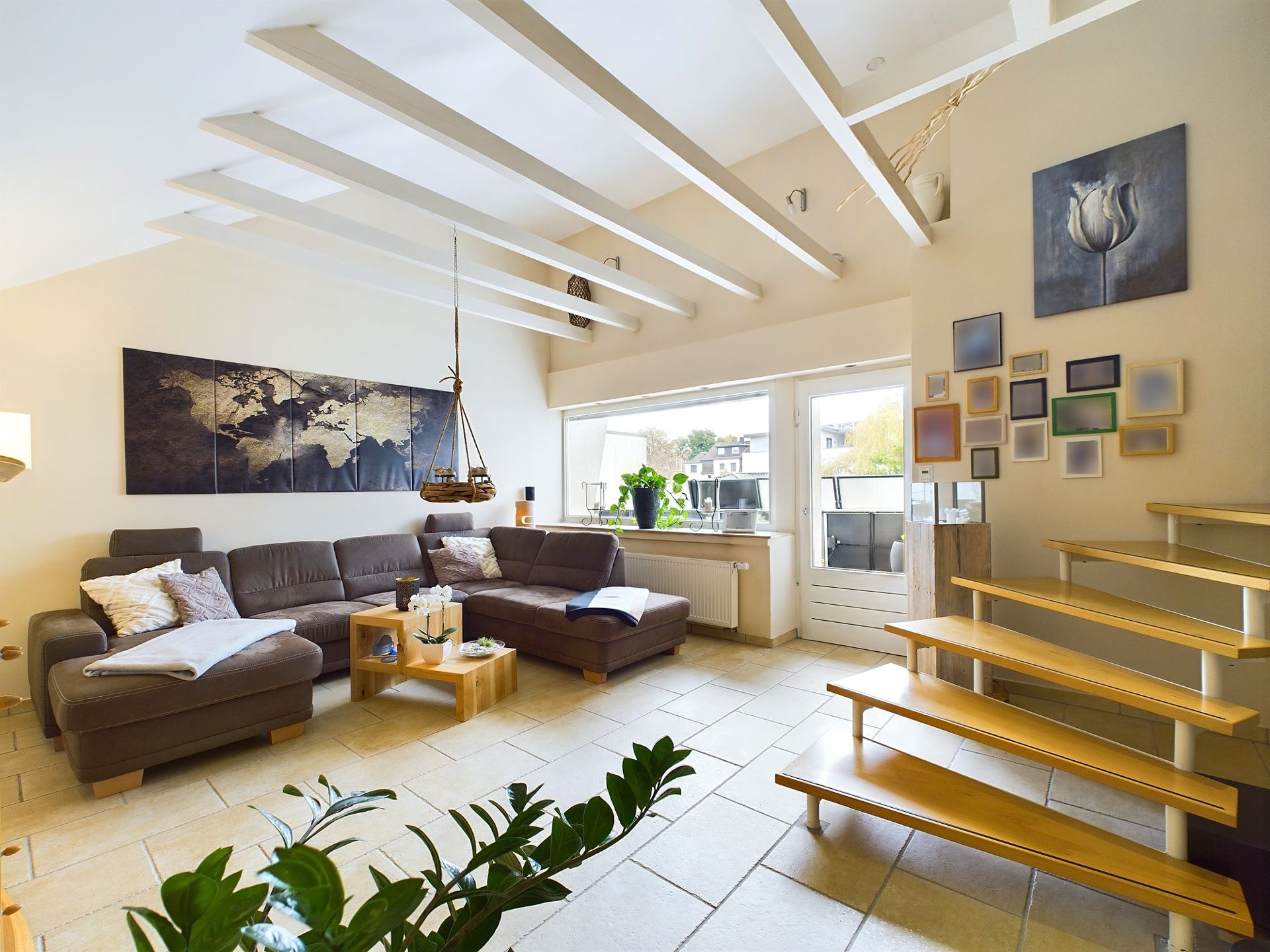Helles Wohnzimmer mit Ecksofa, Wandbildern, Treppe und großen Fenstern.