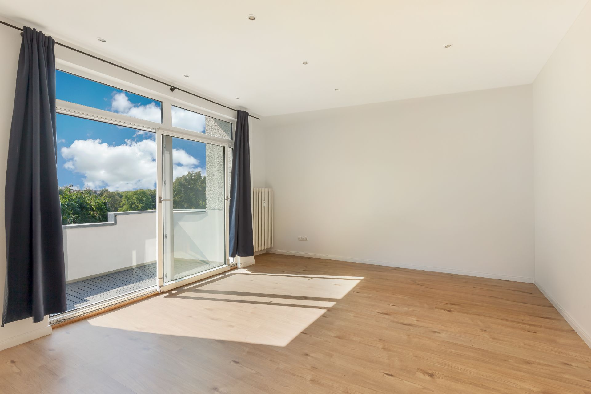 Helles Wohnzimmer mit Holzboden und großen Fenstern, die auf einen Balkon führen.