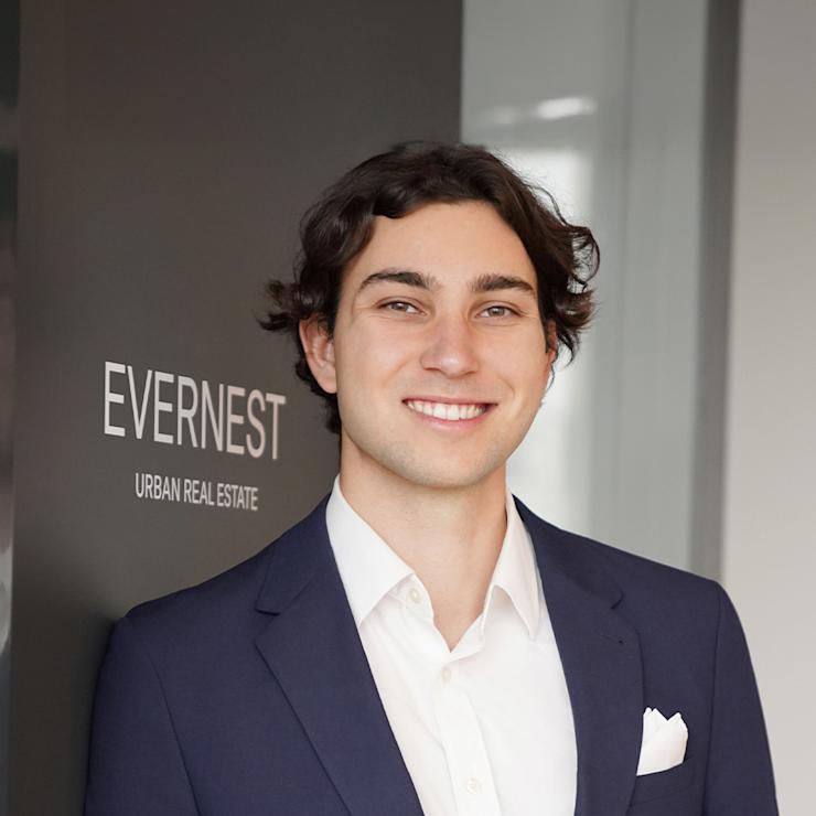 Junger Mann im Anzug steht lächelnd vor einer Wand mit der Aufschrift EVEREST URBAN REAL ESTATE.