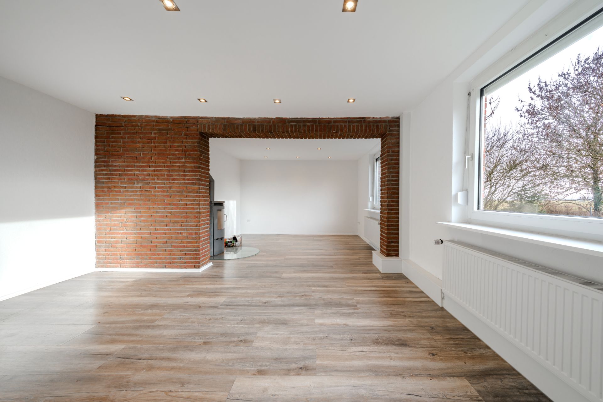 Heller Raum mit Holzboden, großen Fenstern und rotem Backsteinbogen in der Mitte.
