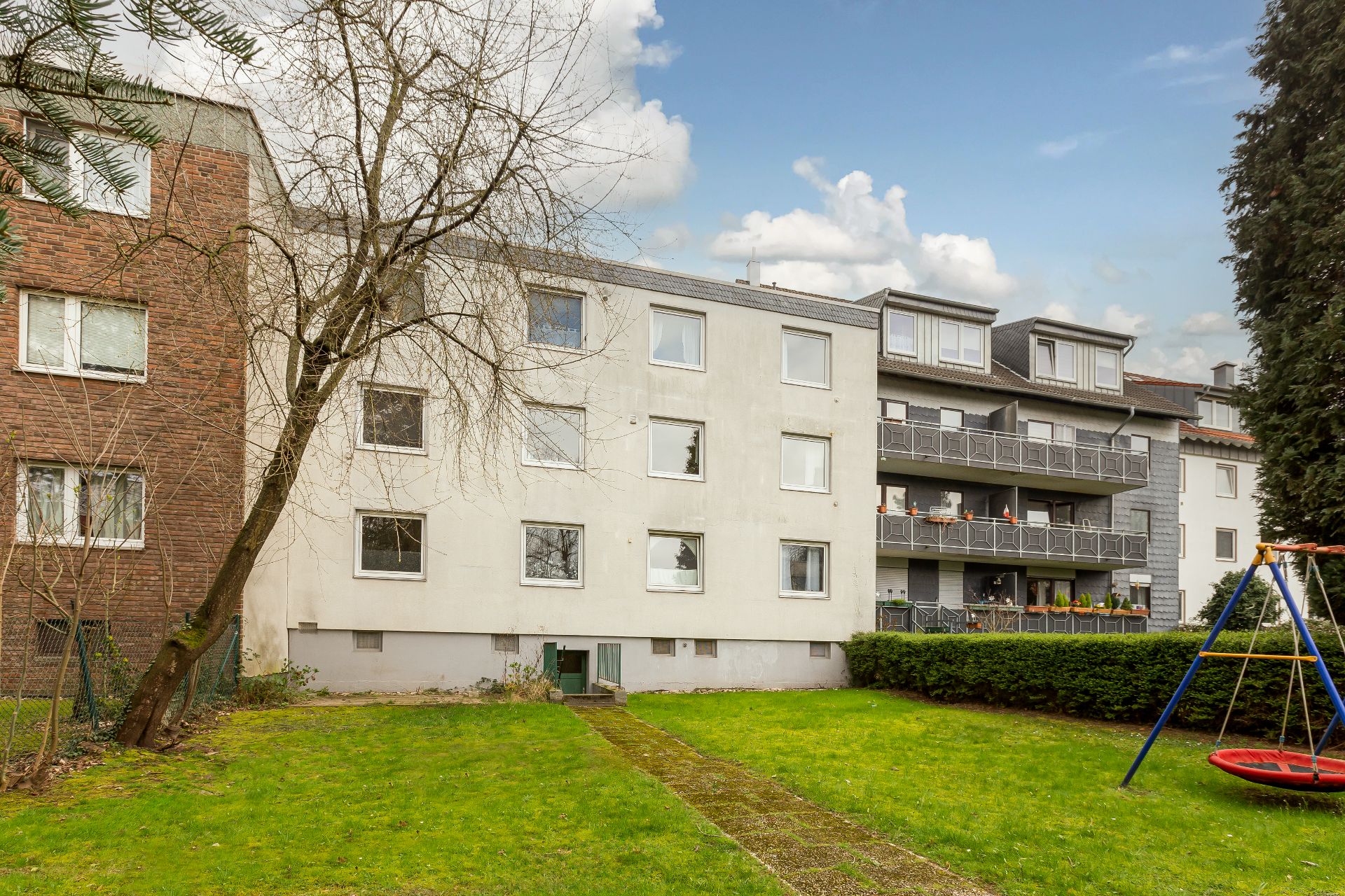 Mehrfamilienhaus mit Garten und Schaukel, im Vordergrund ein Baum und Rasenfläche.