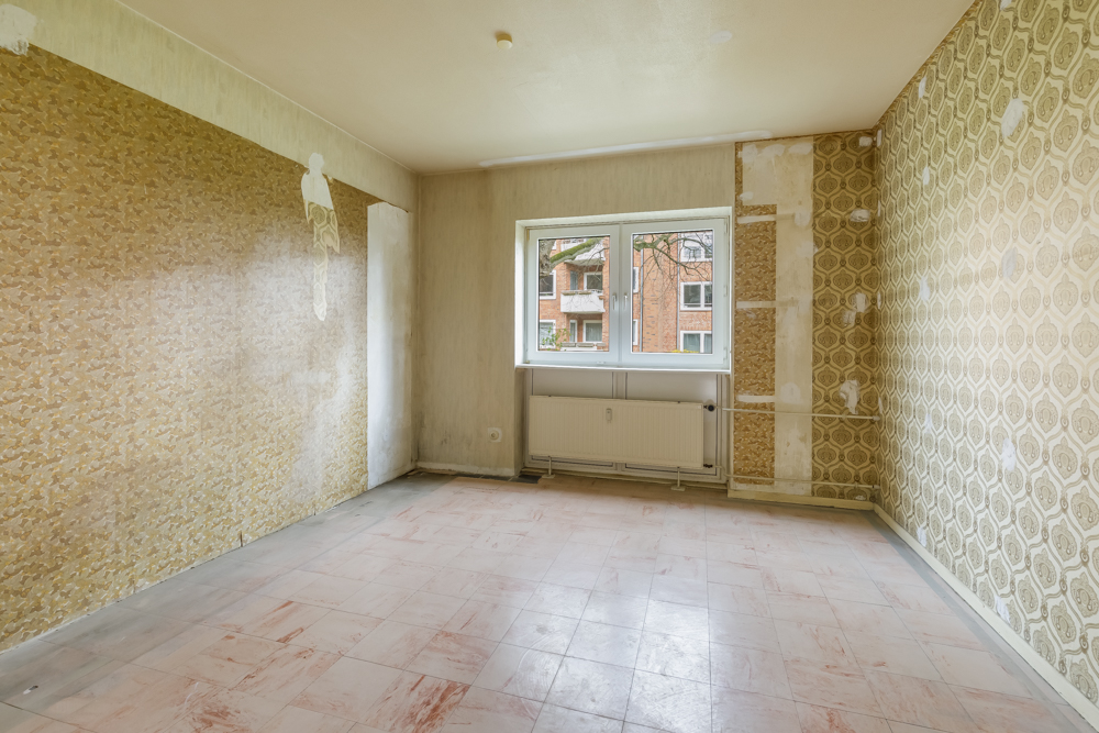 Leerer Raum mit gemusterten Tapeten, großem Fenster und Heizung unter dem Fenster.