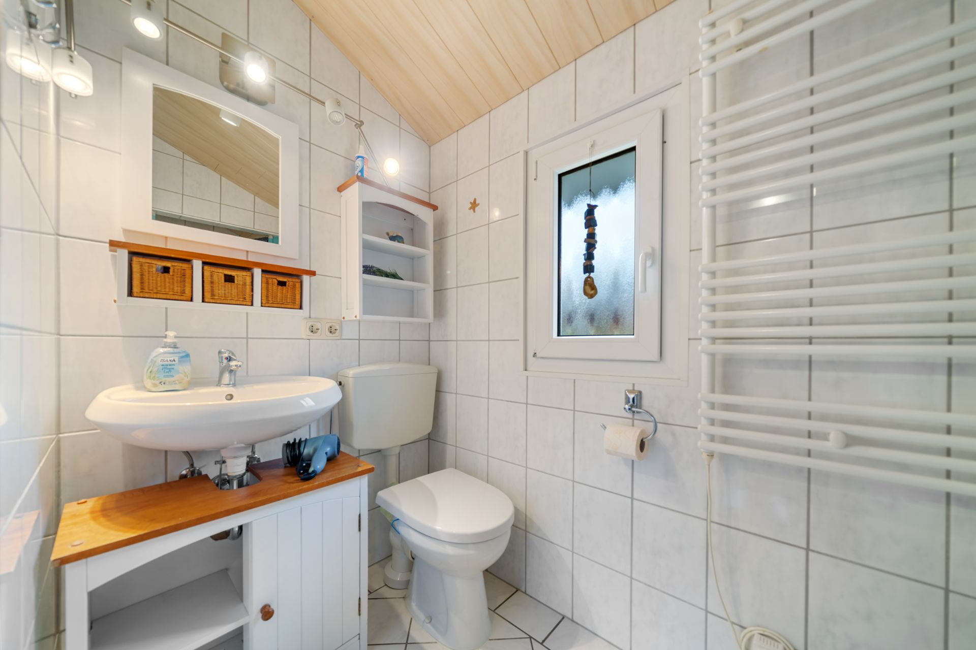Kleines, helles Badezimmer mit Waschbecken, Toilette, Handtuchheizkörper und Fenster.