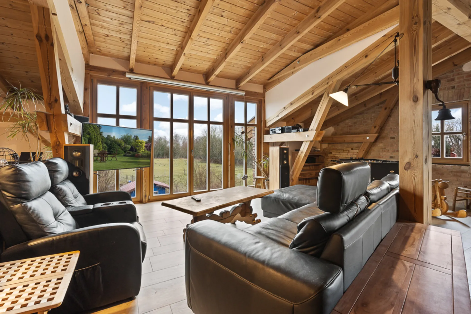Wohnzimmer mit Holzbalkendecke, großen Fenstern, Ledersofas und Blick auf Garten.
