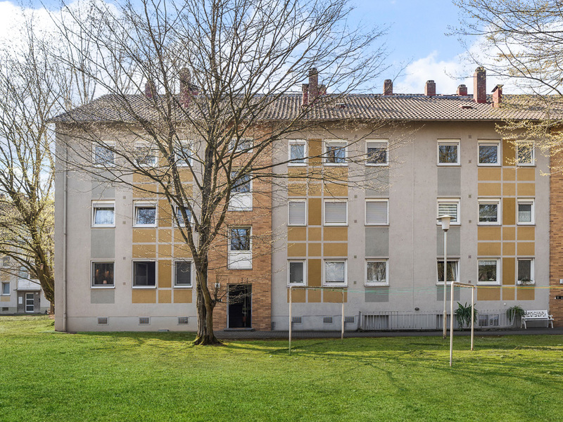 Mehrfamilienhaus mit drei Etagen, großen Fenstern und Vorgarten mit Bäumen.