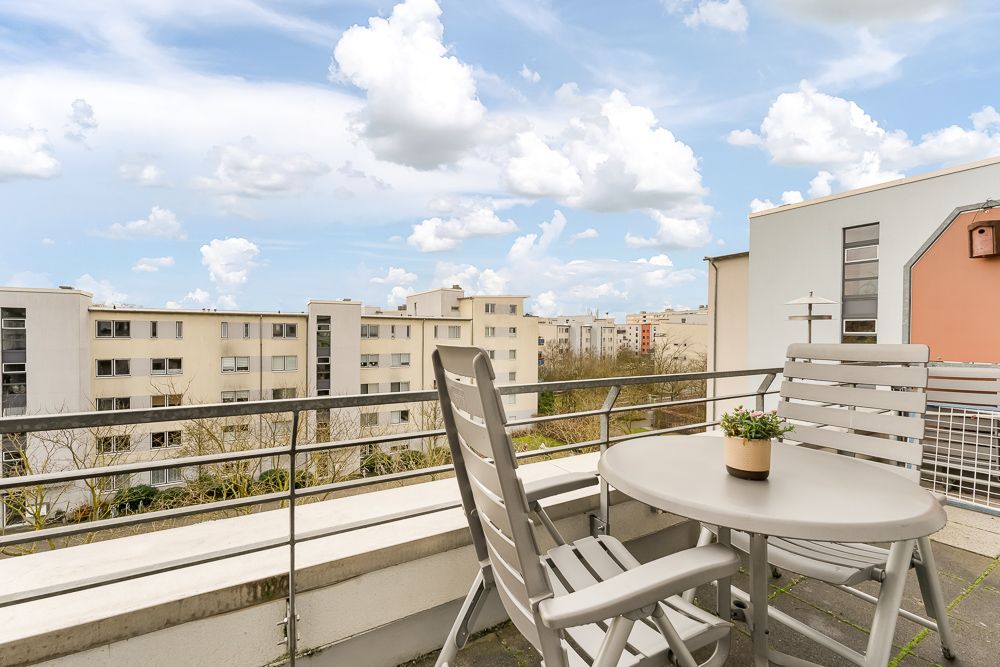 Balkon mit Tisch, zwei Stühlen und Blumentopf, Blick auf Wohngebäude im Hintergrund.
