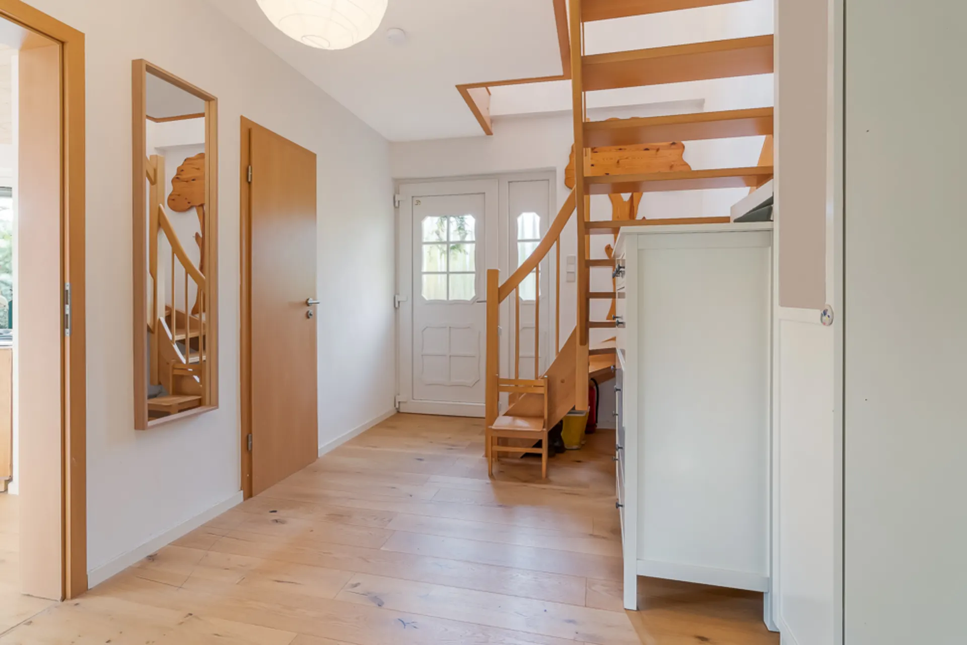 Heller Flur mit Holztreppe, weißer Tür, Spiegel und Holzfußboden.