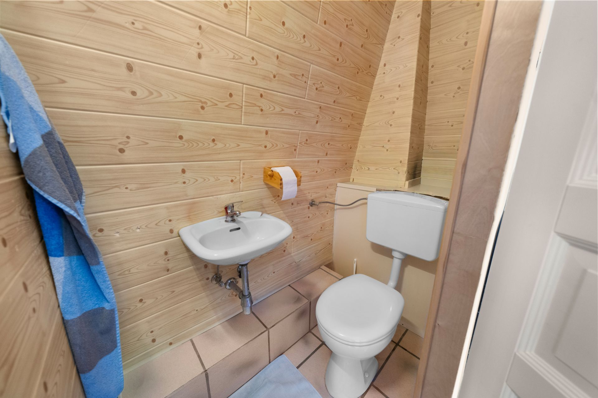 Kleines Bad mit Holzverkleidung, Waschbecken, Toilette, Handtuch und Toilettenpapierhalter.