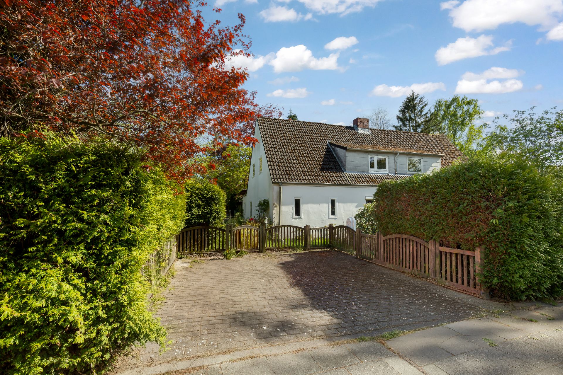 Weißes Wohnhaus mit Garten und kleiner Einfahrt, umgeben von Hecken und Bäumen.