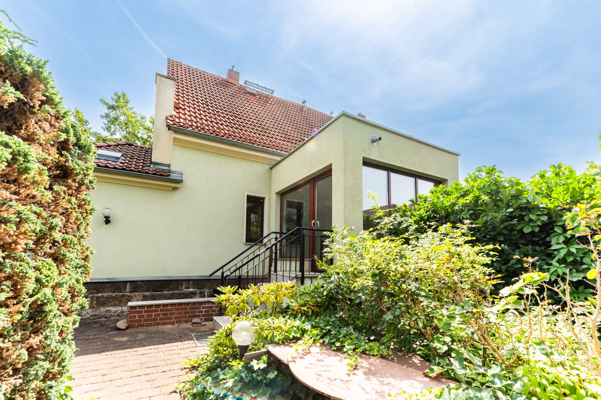 Haus mit gelber Fassade, rotem Dach und Gartentreppe, umgeben von grünen Büschen.