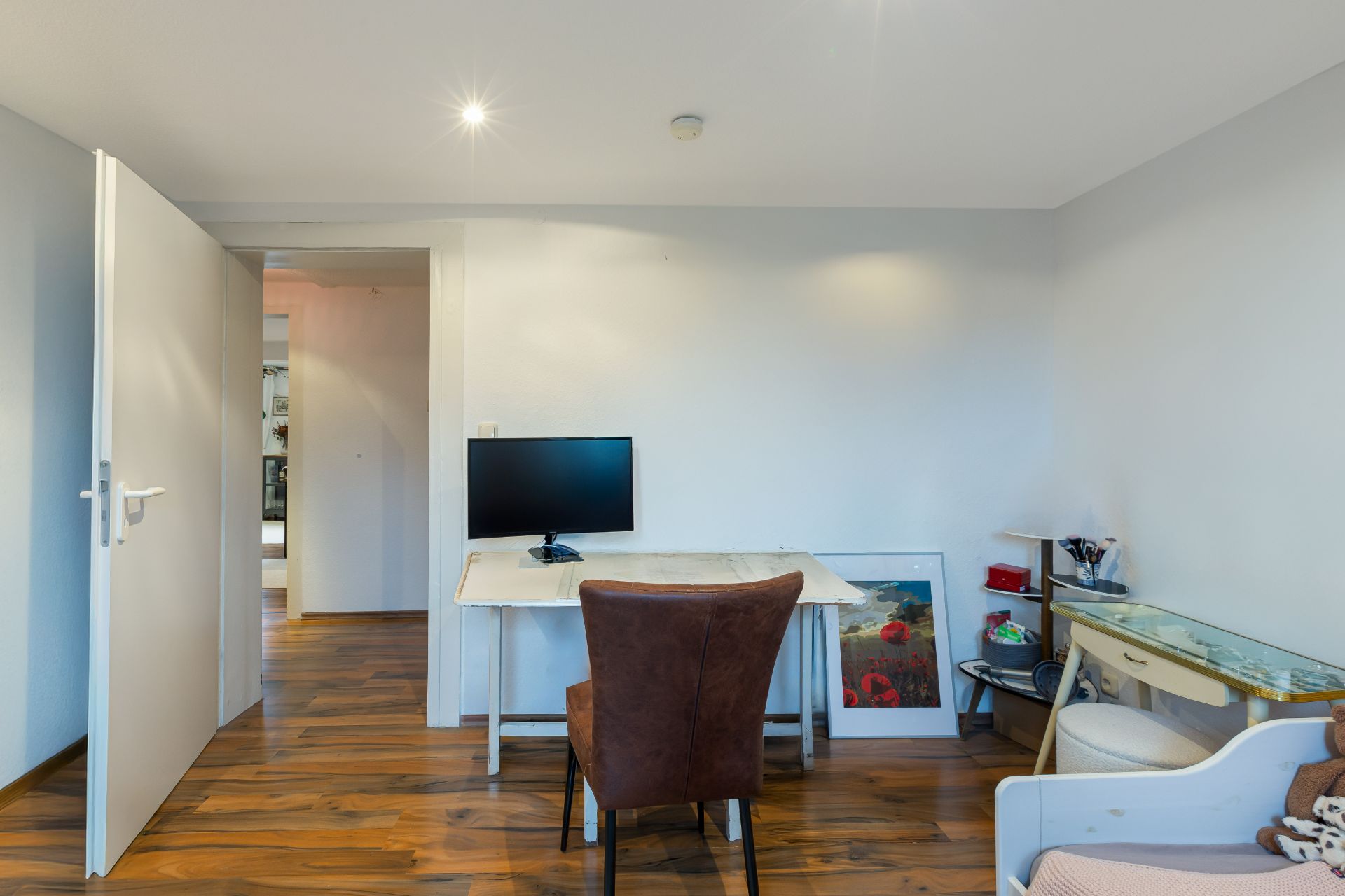 Helles Zimmer mit Schreibtisch, Monitor, braunem Stuhl und Holzfußboden.