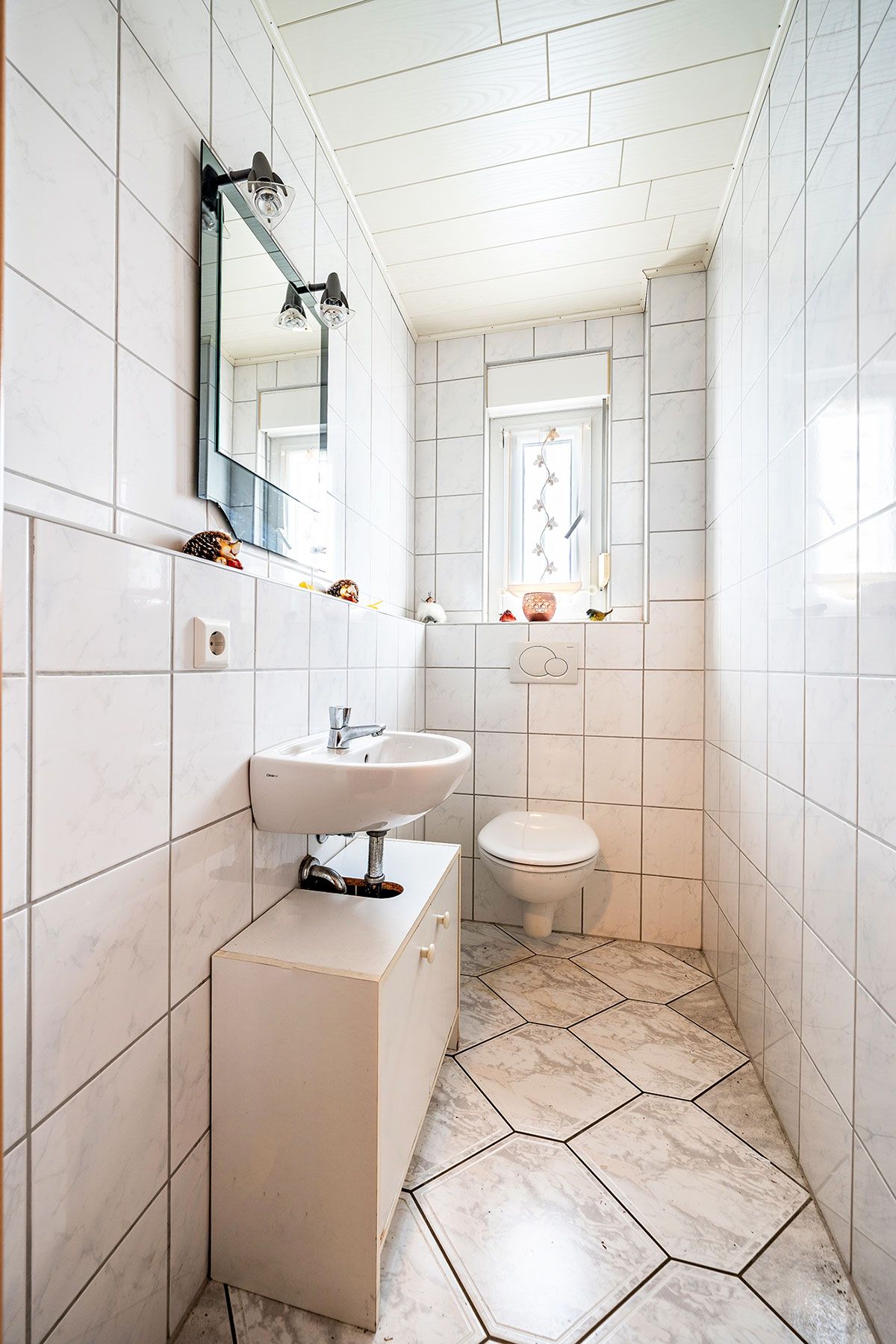 Helles, schmal gefliestes Bad mit Waschbecken, Spiegel, Toilette und Fenster.