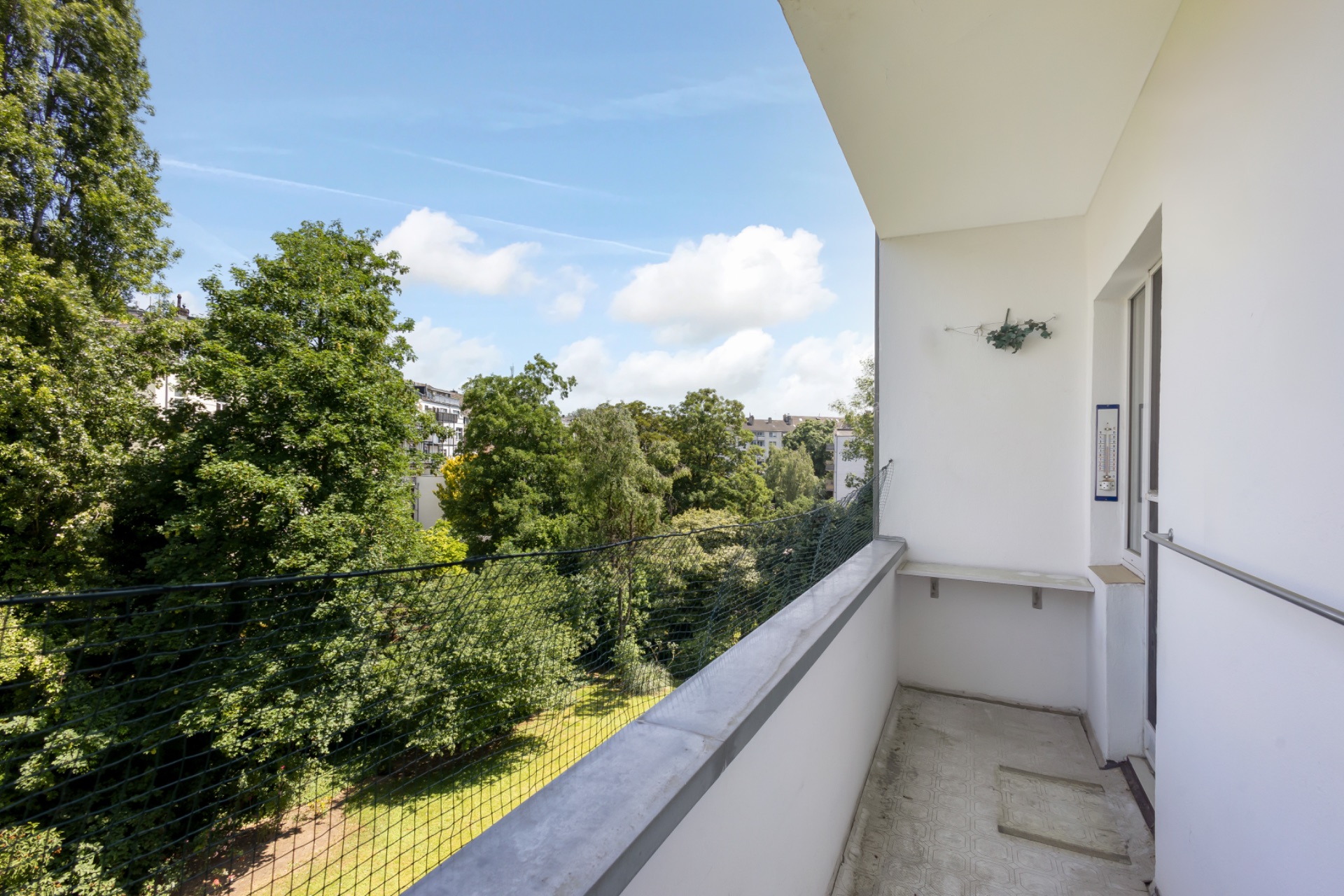 Balkon mit Blick auf grüne Bäume und einen Garten, daneben weiße Hauswand.