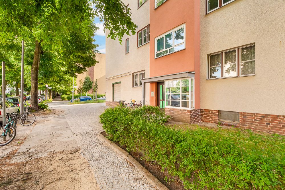Eingang eines Wohnhauses mit großen Fenstern, Gehweg und Büschen davor.