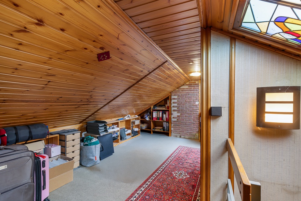 Dachbodenraum mit Holzdecke, Regalen, Teppich und Lagerkisten, beleuchtet durch eine Wandlampe.