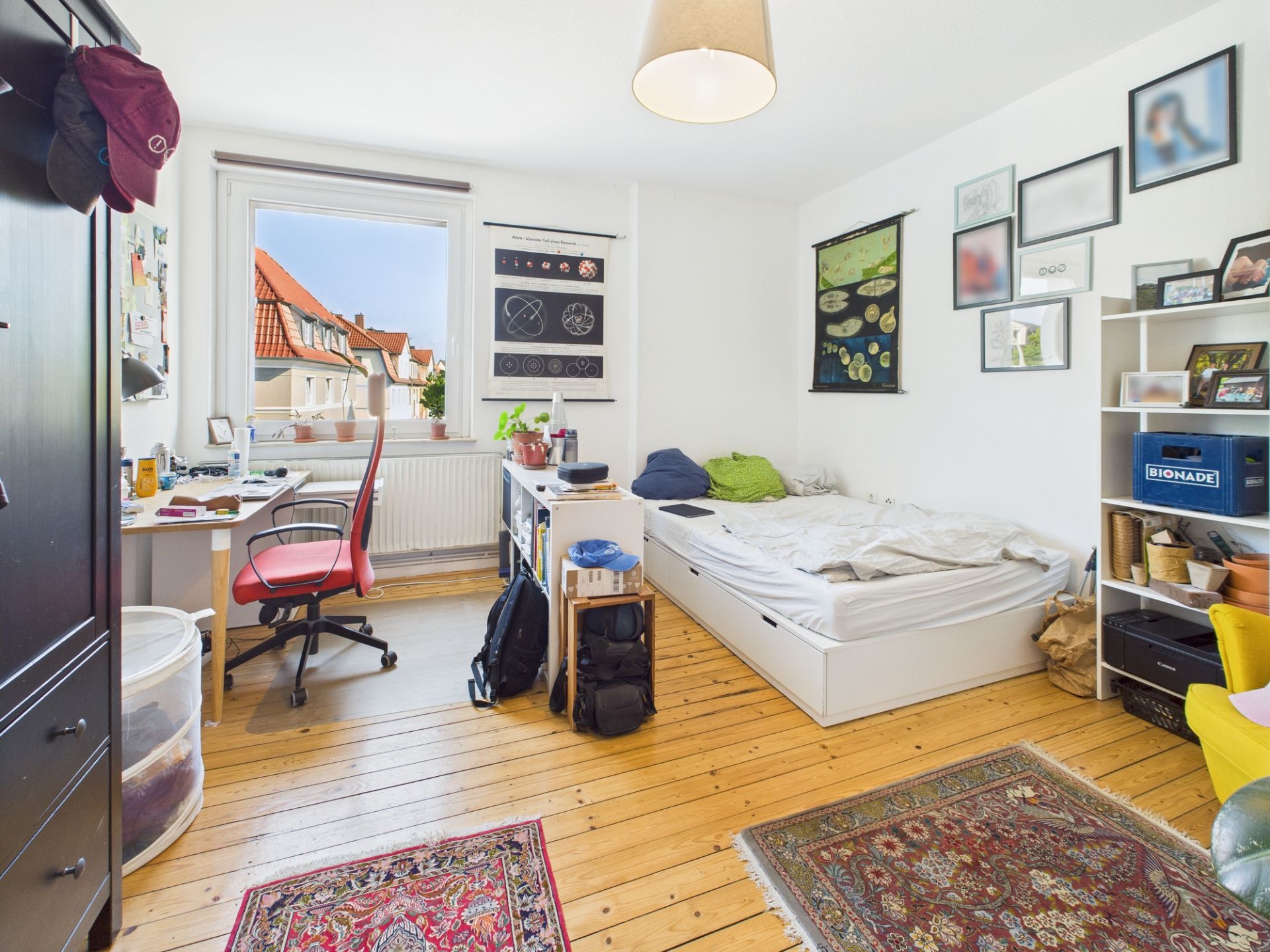 Helles Schlafzimmer mit Holzboden, Bett, Schreibtisch, Regal und Fenster mit Blick nach draußen.