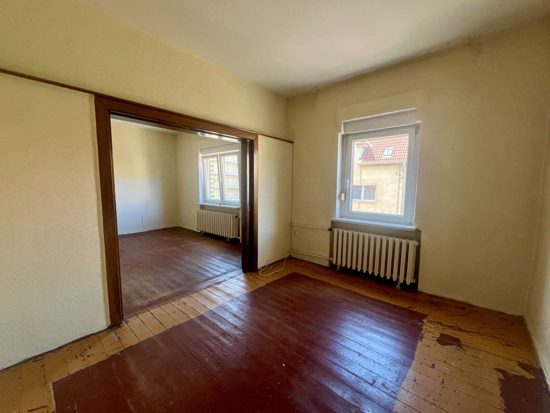 Zwei leere Zimmer mit Holzboden, weißen Wänden und Fenstern mit Heizkörpern darunter.