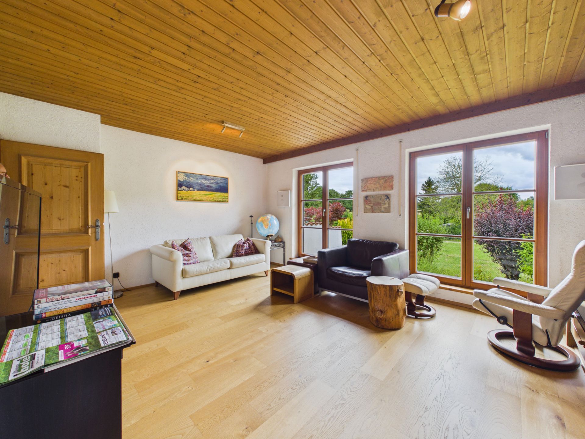Helles Wohnzimmer mit Holzboden, Sofas, Sesseln und großen Fenstern mit Blick auf einen Garten.