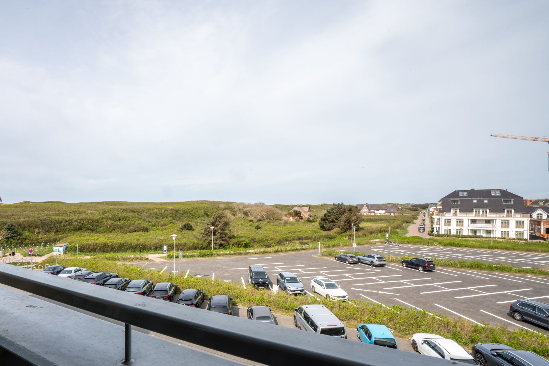 Blick von einem Balkon auf einen Parkplatz, mehrere Autos und eine grüne Landschaft im Hintergrund.