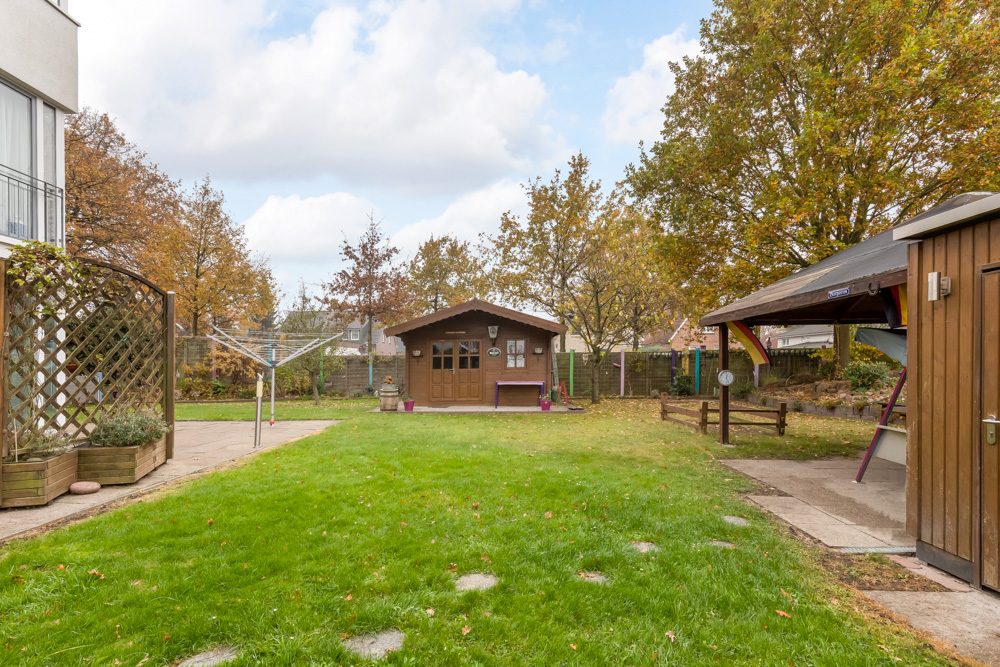 Garten mit Rasen, Holzhaus, überdachtem Bereich und Wäscheleine, umgeben von Bäumen im Herbst.