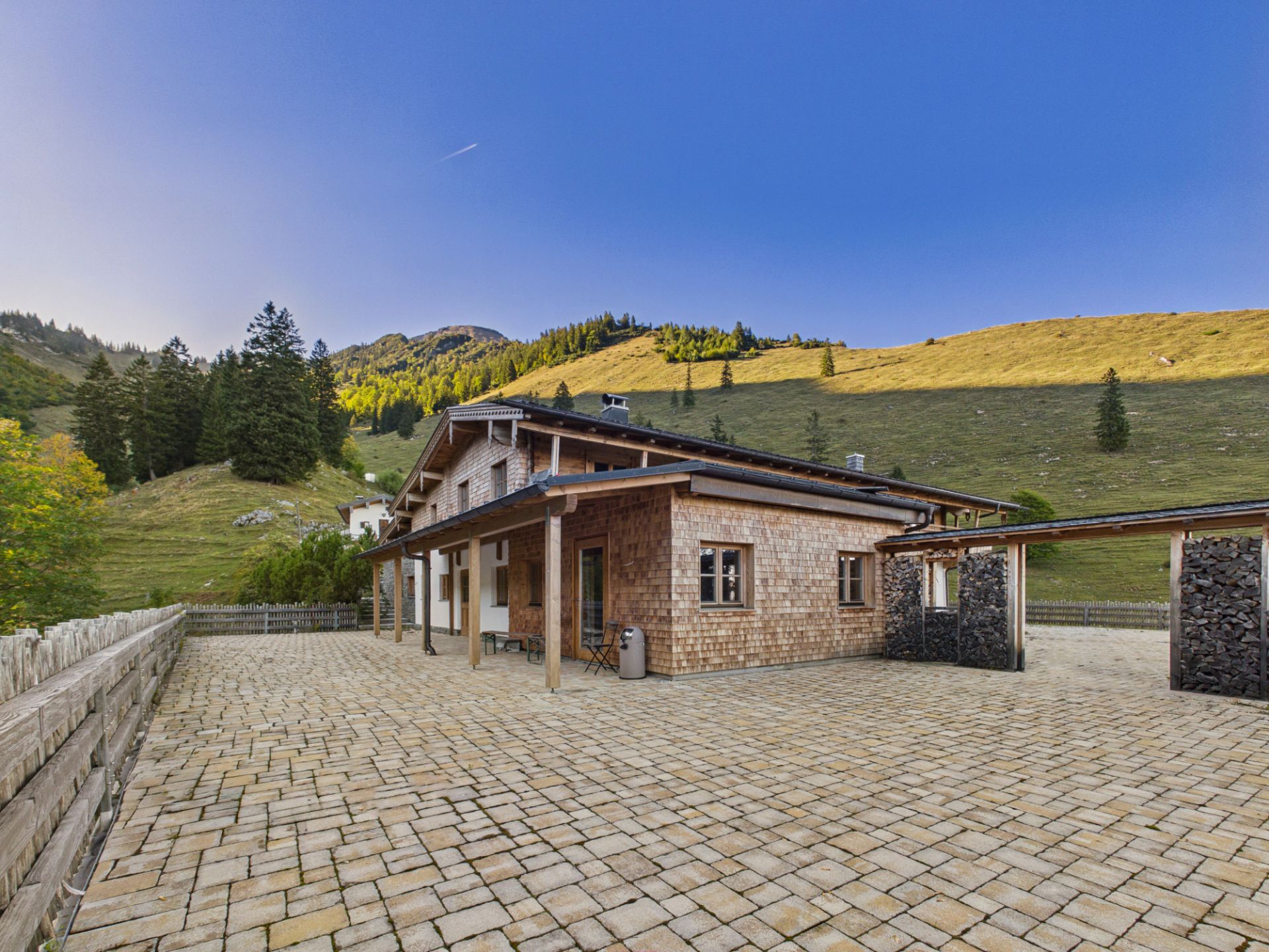Holzhaus mit Terrassenüberdachung vor einer hügeligen Landschaft mit Bäumen und blauem Himmel.