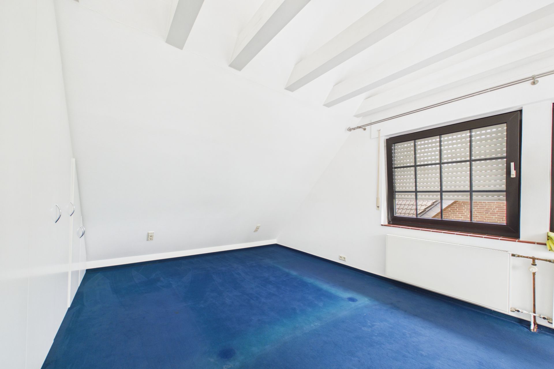Leeres Zimmer mit blauem Teppichboden, weiß gestrichenen Wänden und einem Fenster mit Rollladen.