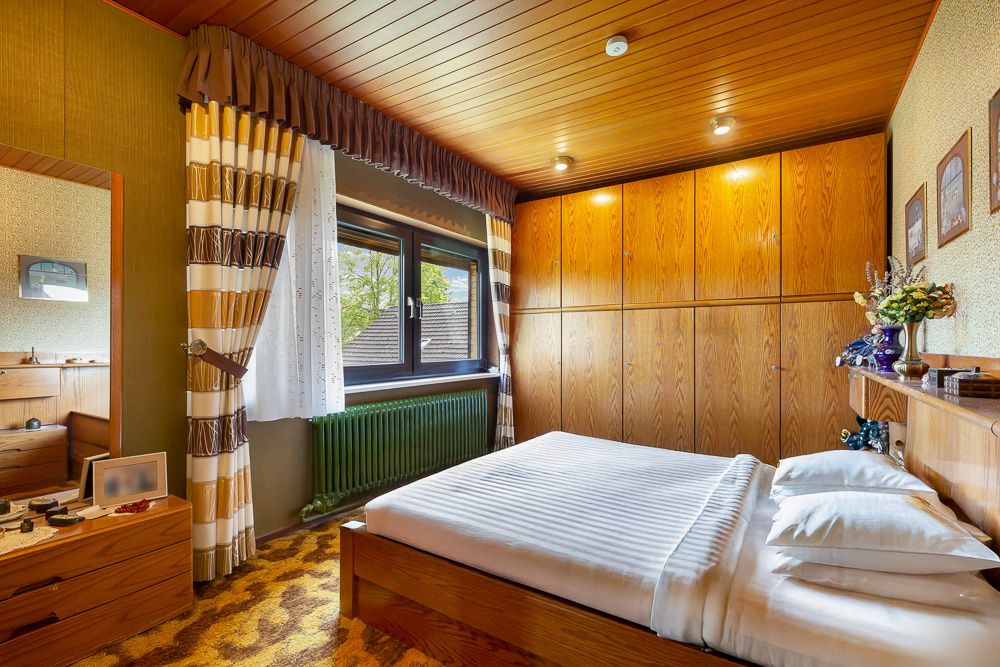 Schlafzimmer mit Holzdecke, Doppelbett, Kleiderschrank, Spiegel und Fenster mit Vorhängen.