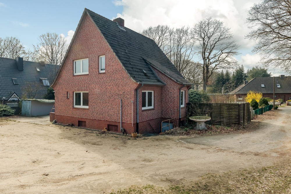 Backsteinwohnhaus mit Spitzdach und mehreren Fenstern, steht auf sandigem Vorplatz mit Bäumen im Hintergrund.