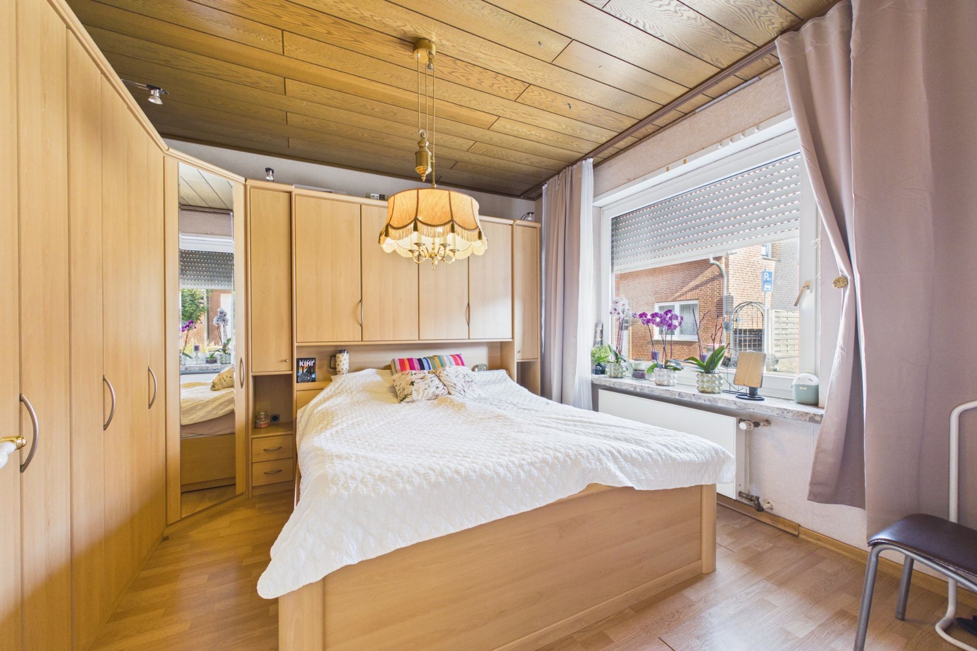Helles Schlafzimmer mit Holzdecke, Doppelbett, Einbauschränken und großem Fenster mit Vorhängen.
