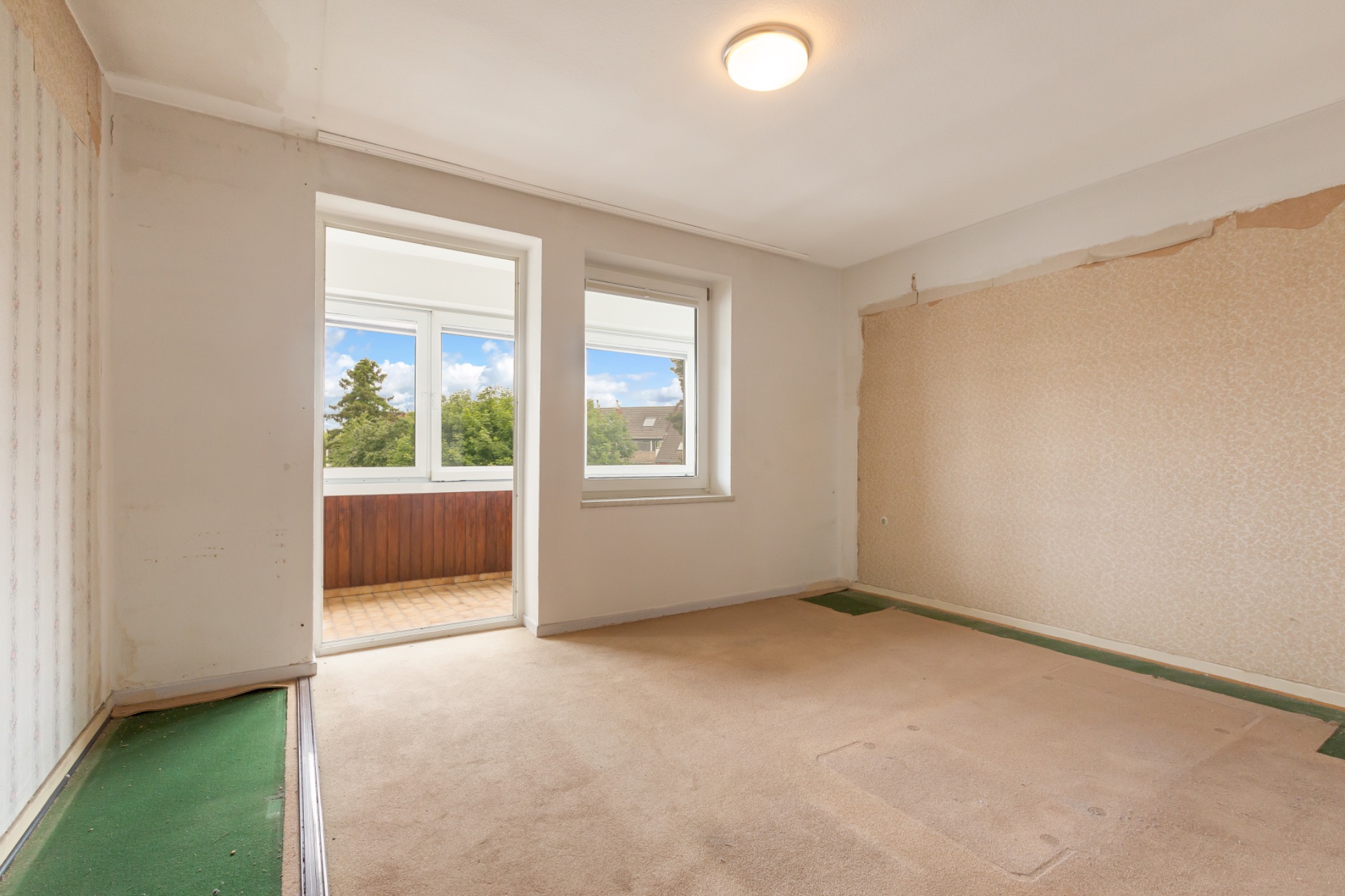 Leeres Zimmer mit beigem Teppich und Zugang zu einer verglasten Loggia, Tageslicht kommt durch große Fenster.