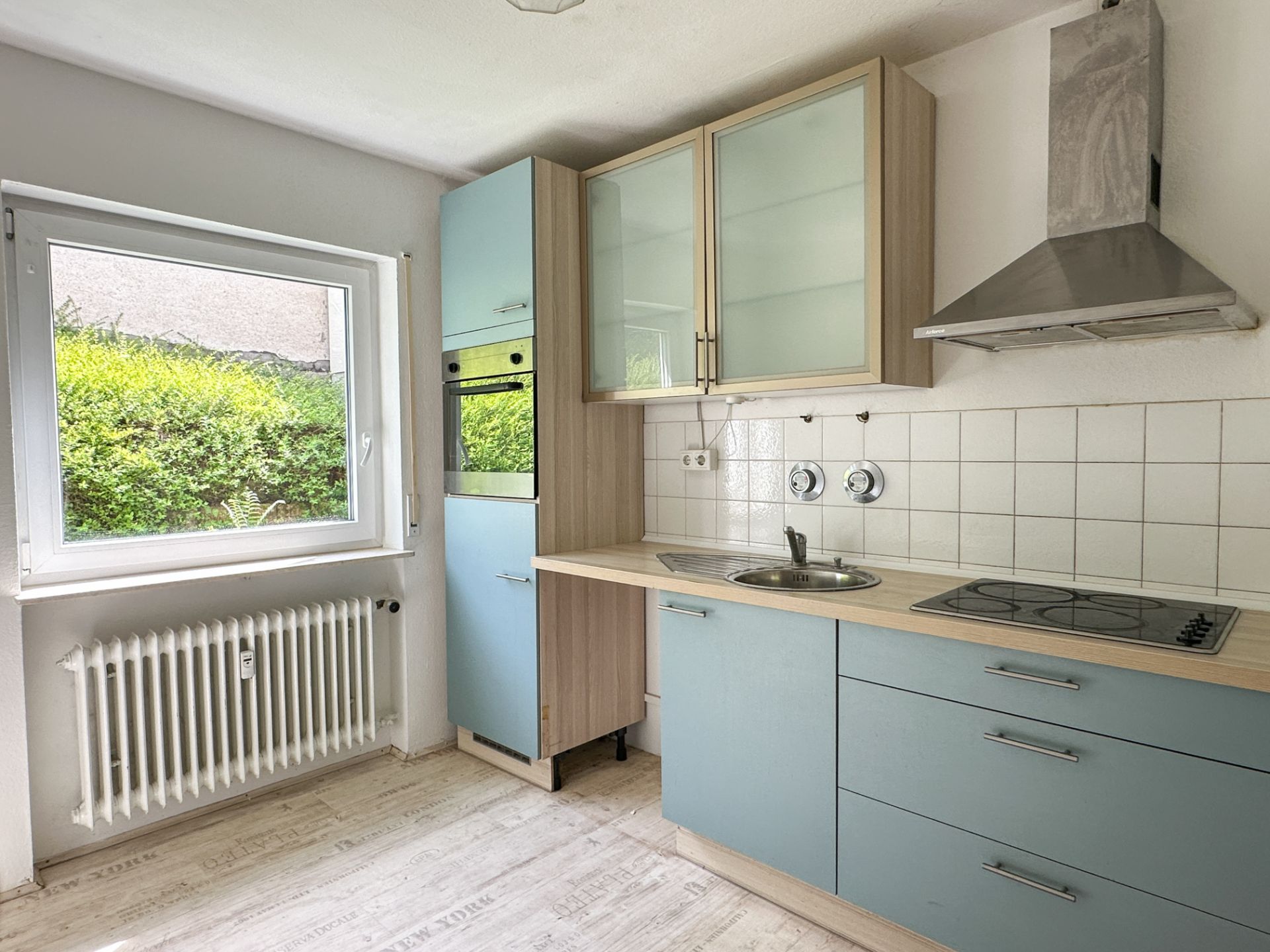 Helle Küche mit blau-grauen Schränken, Fenster, Herd, Spüle und Dunstabzugshaube.