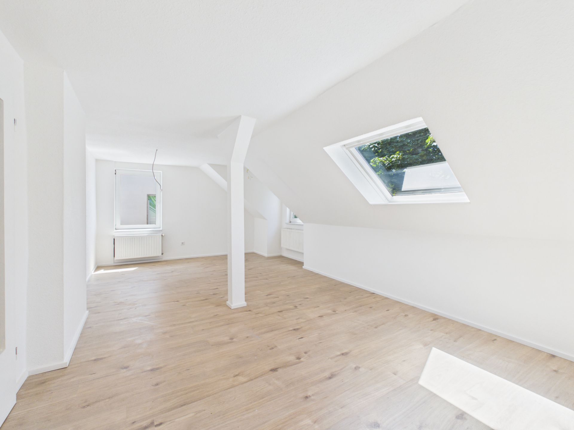 Helles Dachzimmer mit weiß gestrichenen Wänden, Parkettboden und zwei Fenstern.