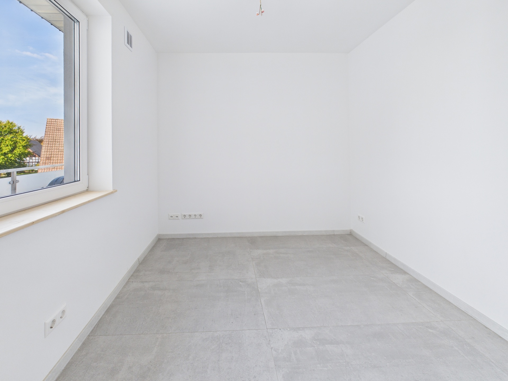 Leerer, heller Raum mit weiß gestrichenen Wänden, großem Fenster und grauem Fliesenboden.