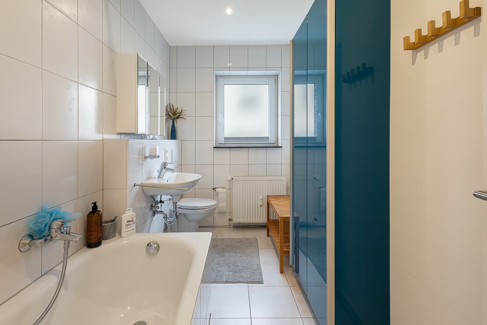 Modernes Badezimmer mit Badewanne, Waschbecken, WC, Fenster und blauem Duschbereich.