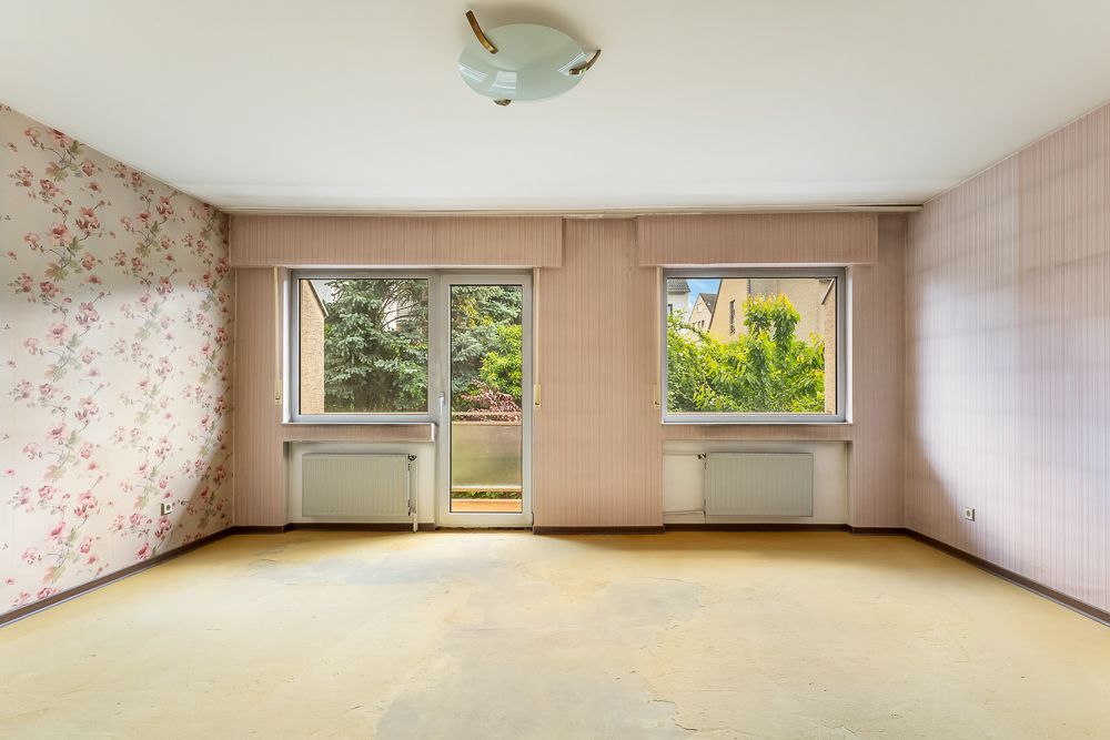 Leerer Raum mit Tapeten, großen Fenstern, Tür zum Balkon und Heizkörpern unter den Fenstern.