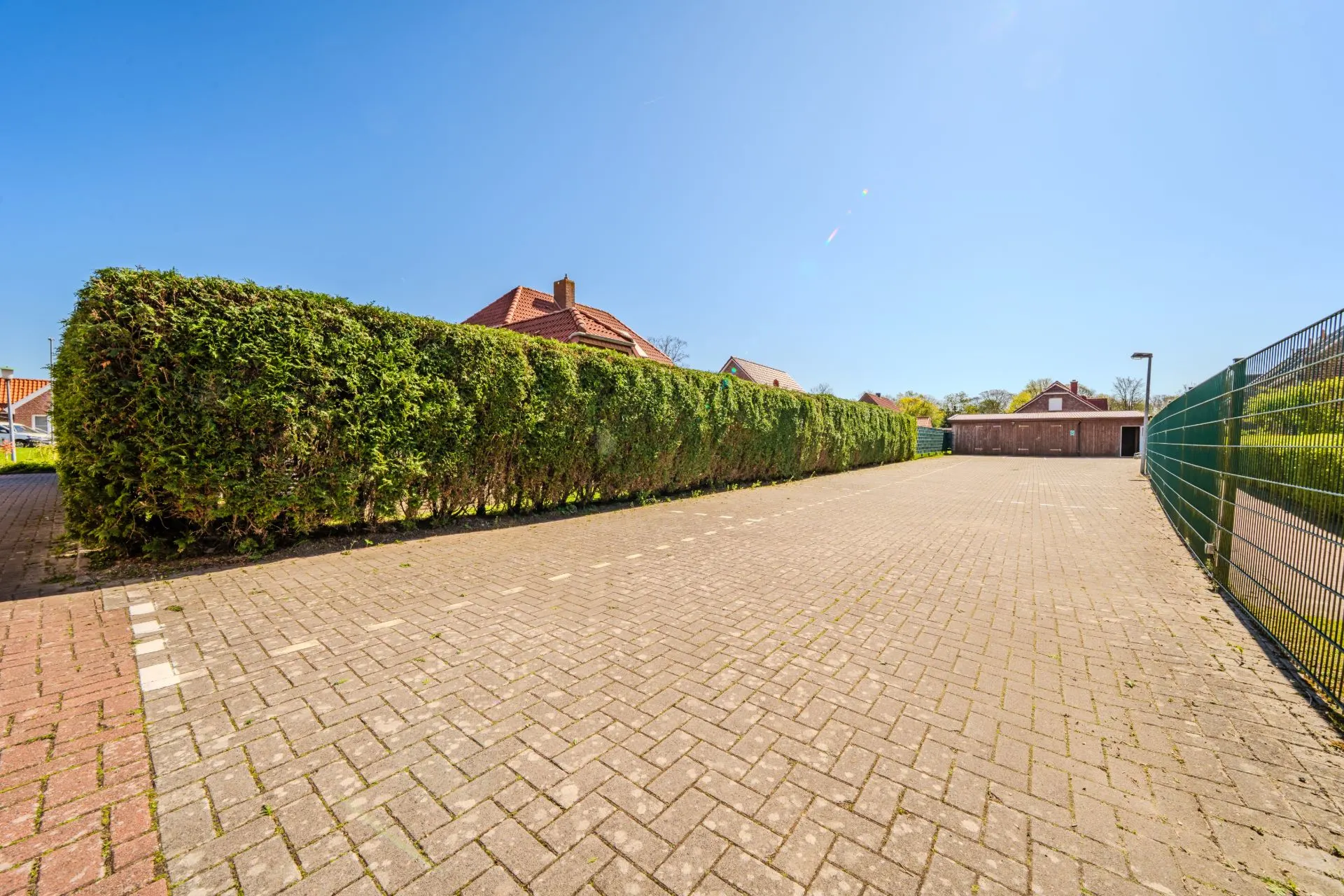 Gepflasterter Parkplatz mit Hecke und Sicht auf Wohnhäuser im Hintergrund.