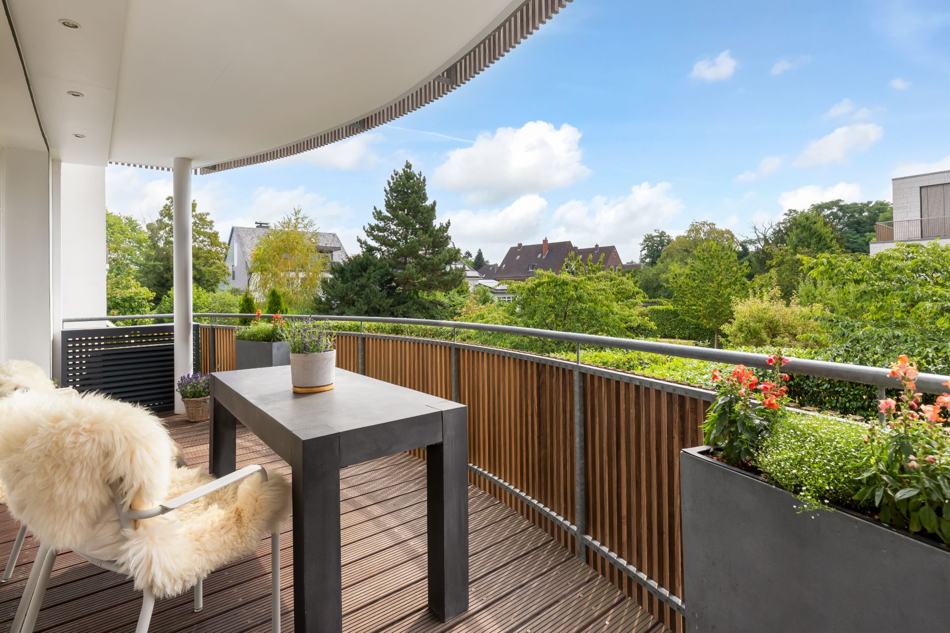 Moderner Balkon mit Holzboden, Tisch, Stuhl mit Fellauflage und Pflanzen, Blick ins Grüne.