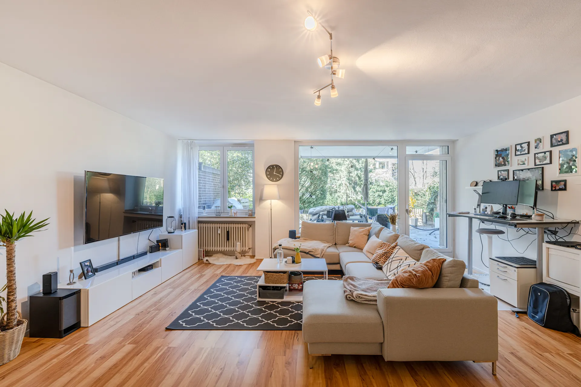 Helles Wohnzimmer mit Ecksofa, Fernseher, Teppich und Schreibtisch am Fenster.