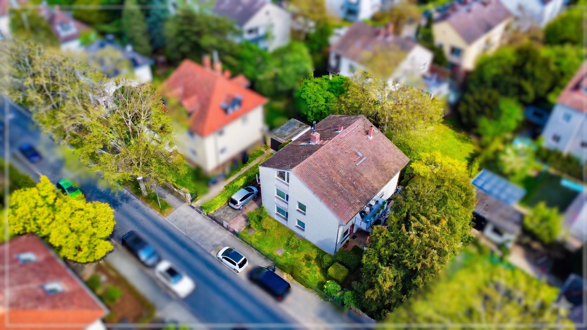 Luftaufnahme eines weißen Wohnhauses mit Garten an einer Straße, umgeben von Bäumen und weiteren Häusern.