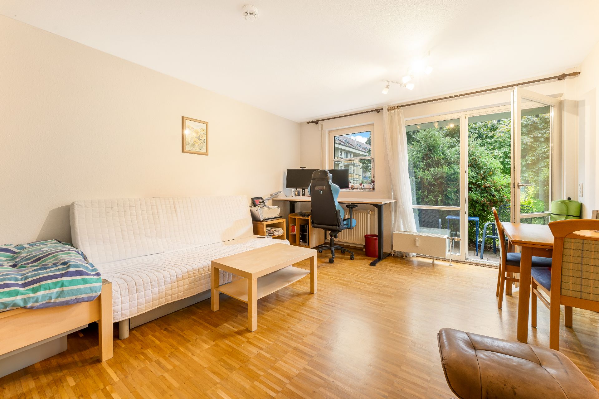 Helles Wohnzimmer mit Sofa, Schreibtisch, Essbereich und Zugang zu einem Balkon mit Blick ins Grüne.