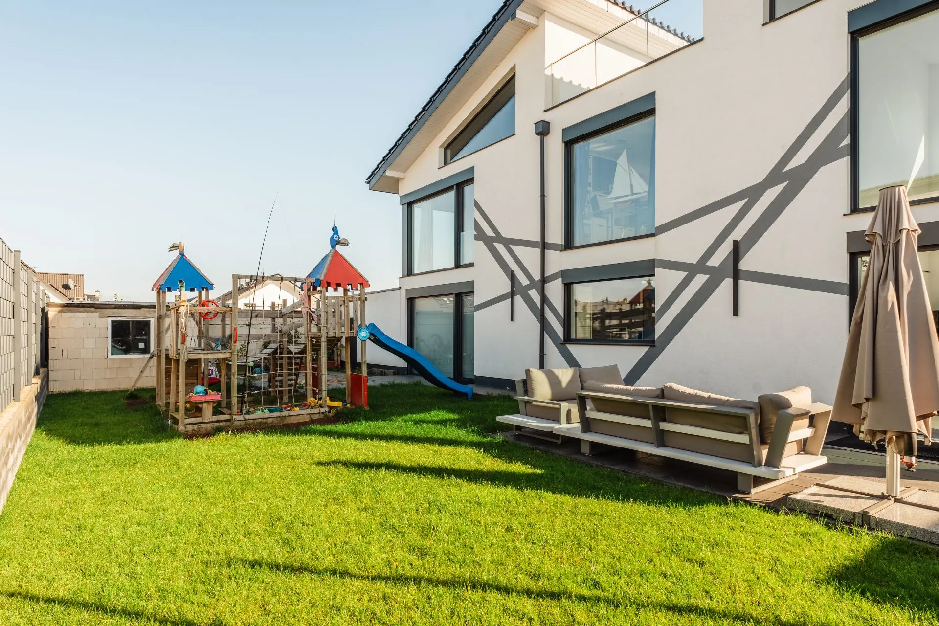 Garten mit Spielplatz, blauer Rutsche, Sitzgruppe und Sonnenschirm neben modernem Haus.