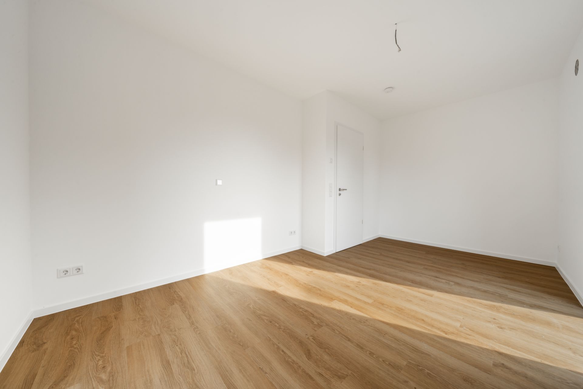 Leerer, heller Raum mit Holzboden, weißen Wänden und einer geschlossenen Tür.