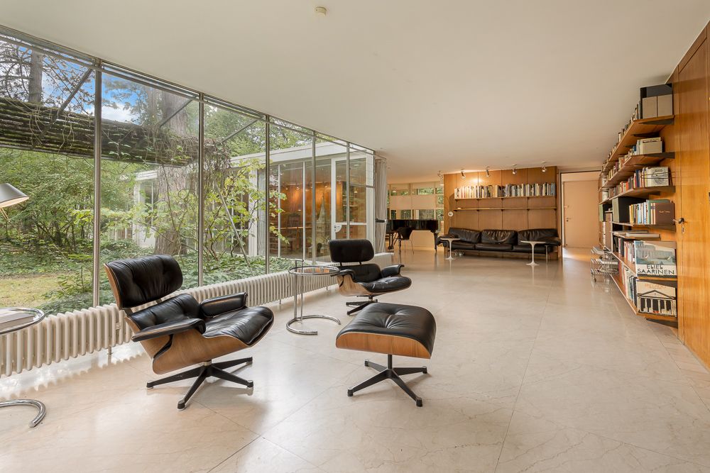 Modernes Wohnzimmer mit großen Fenstern, schwarzen Sesseln, Regalwand und Blick ins Grüne.